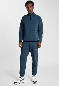 Granatowy sportowy dres wykonany z gładkiej tkaniny; bluza z zamkiem z raglanowymi rękawami oraz zwężane spodnie z bocznymi kieszeniami i detalem z logo.