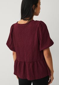Bordeaux-farvet bluse med struktur, korte pufærmer og peplum-kant. Har en krøllet stoffinish og en rund halsudskæring.