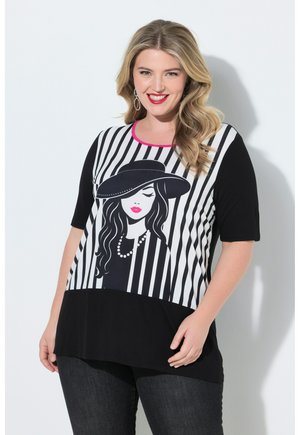 Smilende kvinde med bølget blondt hår iført sorte jeans og en sort-hvidstribet top med et grafik af en kvinde med hat og pink læber.