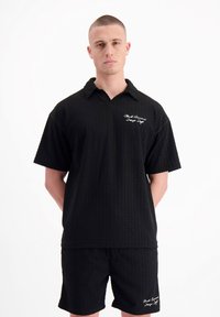 Zwart geweven shirt en short set met een textuurpatroon. Heeft een kraag en een geborduurd logo op de linkerborst.