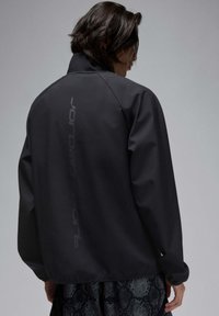 Chaqueta negra, ligera, con un alto cuello y un sutil logo embosado verticalmente en la parte posterior; textura suave; combinada con una prenda inferior de diseño estampado.