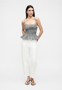 Mujer con top sin mangas de cuadros en blanco y negro con volante en el dobladillo, pantalones blancos de pierna ancha y sandalias negras tipo tanga, de pie con las manos detrás de la espalda.