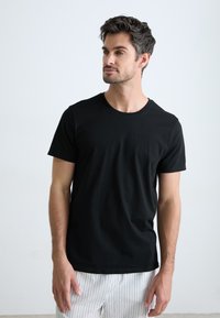 T-shirt noir à manches courtes en coton avec un col rond, texture lisse, porté avec un pantalon de pyjama rayé, présentant un design simple.