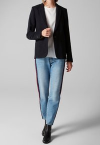 Blazer bleu marine avec deux boutons, associé à un jean bleu clair avec des bandes latérales rouges et des bottines noires. Tissu lisse, coupe ajustée.