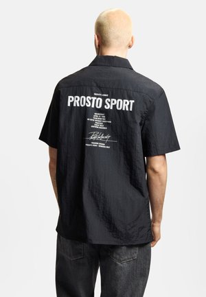 Osoba z krótkimi blond włosami, ubrana w czarną koszulkę z krótkim rękawem z napisem "PROSTO SPORT" i tekstem nadrukowanym na plecach.