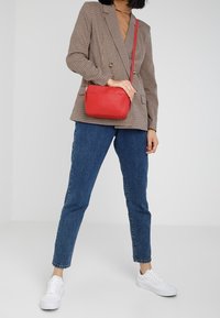 Punainen nahkainen olkalaukku, jossa on sileä pinta, suorakulmainen muoto ja ohut säädettävä hihna. Yhdistetty ruudulliseen bleiseriin ja farkkuihin.