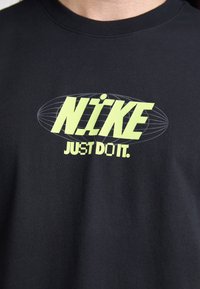 Musta t-paita, jossa on keltainen "Nike" -teksti ja "Just Do It" -slogan ovaalimaisessa langankehysdesignissä.