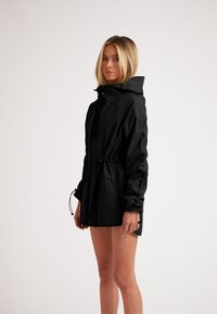 Giacca leggera nera con cappuccio, cintura con cordino e polsini elasticizzati. Texture liscia, design minimalista, che mette in mostra una silhouette elegante e moderna.