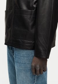 Δεξί χέρι ακουμπισμένο στο πλάι, φορώντας μαύρο δερμάτινο μπουφάν με μπροστινή τσέπη και μπλε τζιν παντελόνι.