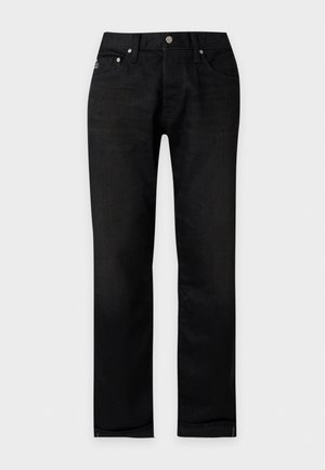 Jeune homme debout portant un t-shirt blanc uni à manches courtes, un pantalon noir et des chaussures noires sur un fond blanc uni.