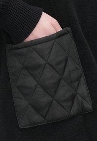 Mustast tikandiga tasku teemantmustriga, kinnitatud tumedale kangale, osaliselt nähtava soonikkrae mansetiga, taskus käsi sees.