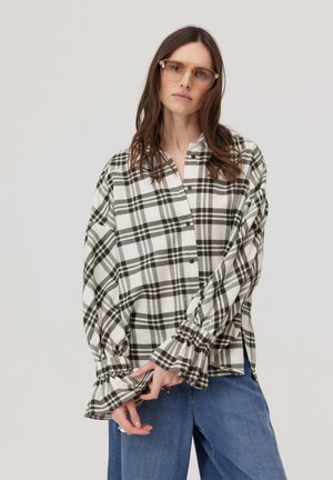 Kobieta ubrana w oversize czarno-białą koszulę w kratę z marszczonymi mankietami i niebieskie dżinsy, pozująca na jednolitym jasnym tle.