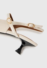 Beige patent leather slingback-kengät, joissa on kaksinkertaiset kultaiset soljet ja musta lievästi kaareva kitten-korko valkoista taustaa vasten.