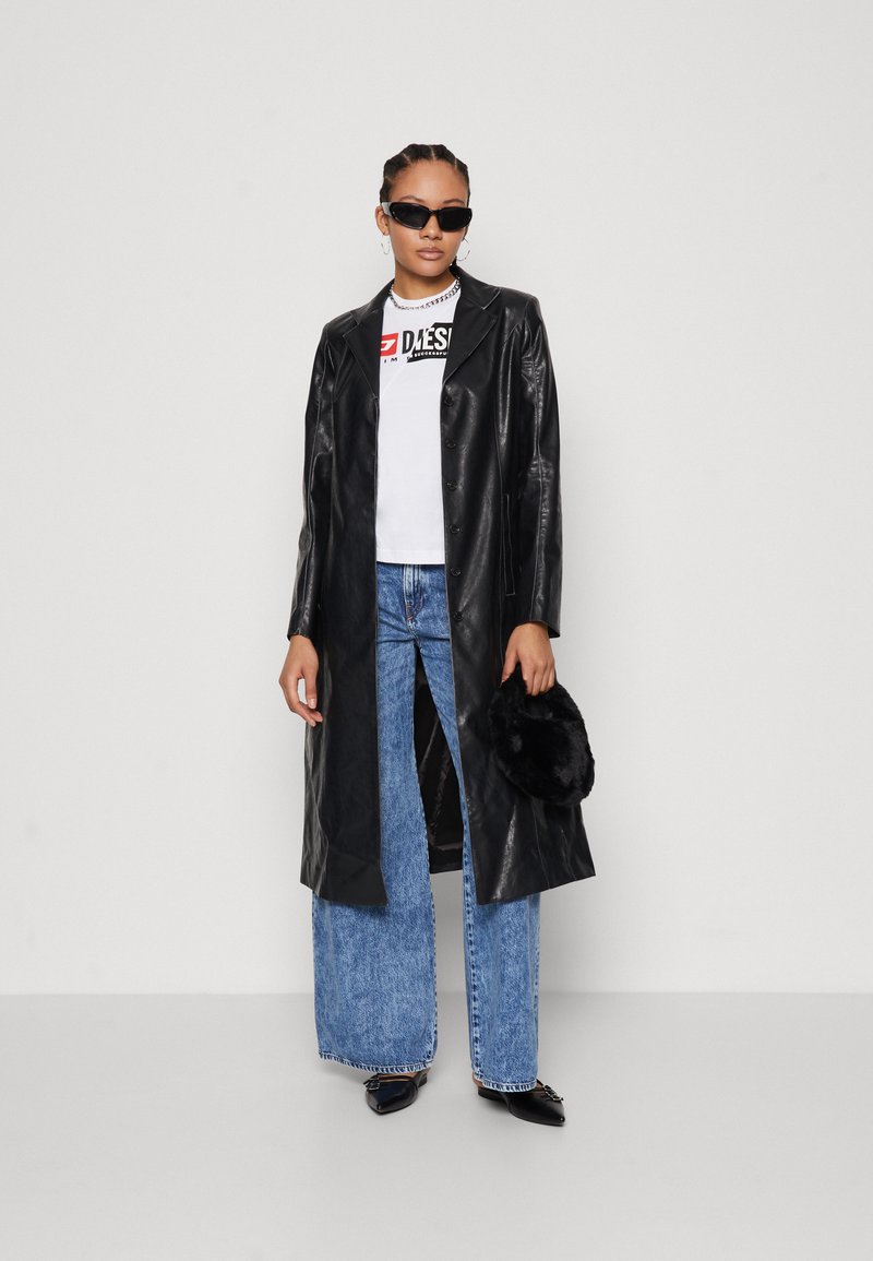Abrigo largo de piel negra con botones, combinado con una camiseta blanca con logo y jeans de mezclilla azul de pierna ancha. Los accesorios incluyen gafas de sol negras y un bolso de pelaje.
