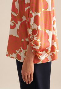 Blusa floreale con sfondo bianco; presenta audaci motivi rosa e oro. Maniche a tre quarti con dettagli arricciati ai polsini.