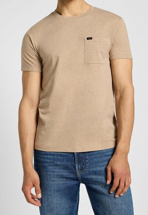 Homme portant un t-shirt beige à manches courtes avec col rond et poche poitrine, associé à un jean en denim bleu, sur un fond uni.