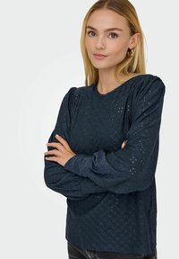 Granatowa bluzka z długim rękawem z wzorami z dziurek, okrągłym dekoltem i lekko bufiastymi ramionami. Gładka faktura z misternymi detalami.