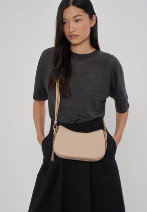 Kobieta z długimi ciemnymi włosami, ubrana w ciemnoszary top z krótkim rękawem, czarną spódnicę oraz beżową torbę na ramię z zamkiem błyskawicznym i paskiem.