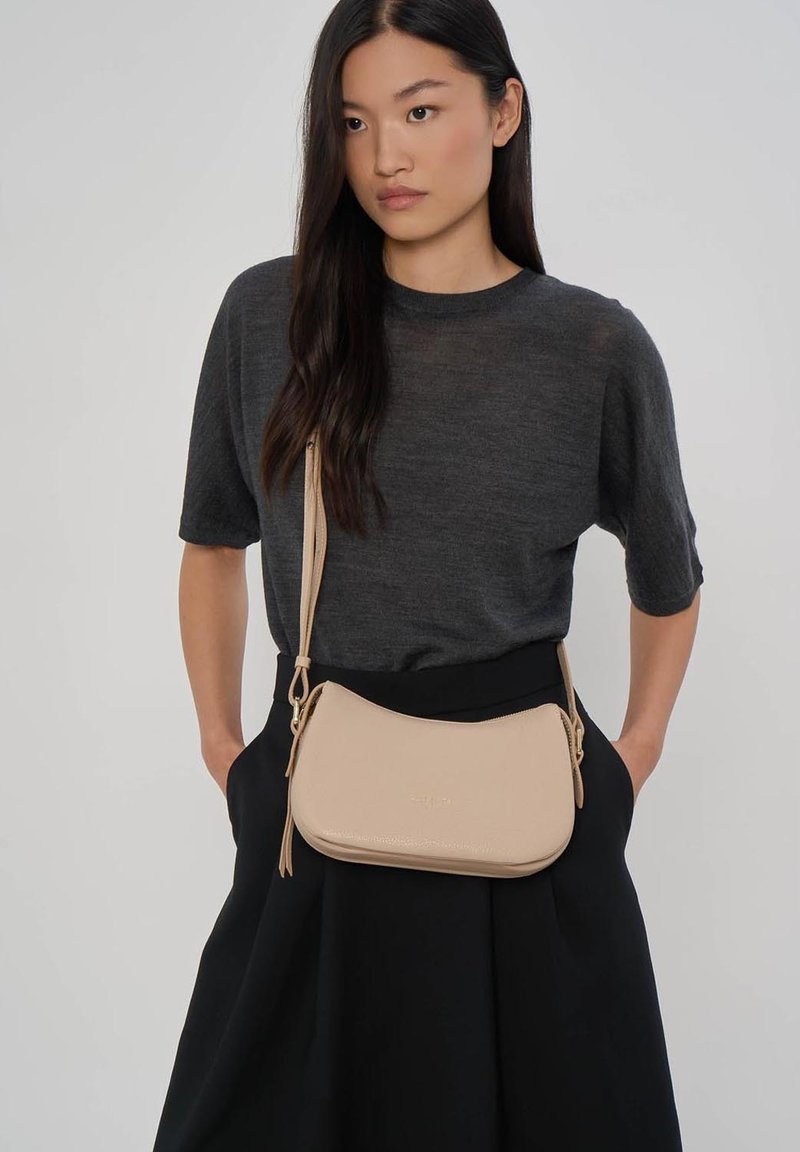 Femme aux longs cheveux noirs portant un haut gris foncé à manches courtes, une jupe noire et un sac bandoulière beige avec fermeture éclair et sangle.