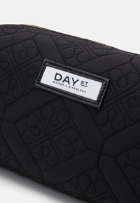 Funda de tela negra acolchada con una etiqueta blanca con la marca "DAY ET". El diseño incluye patrones en relieve y un detalle de cremallera dorada.