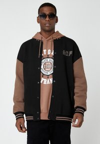 Chaqueta varsity negra y marrón con botones de presión, puños a rayas y bolsillos laterales sobre un sudadera beige con texto gráfico y estrellas.