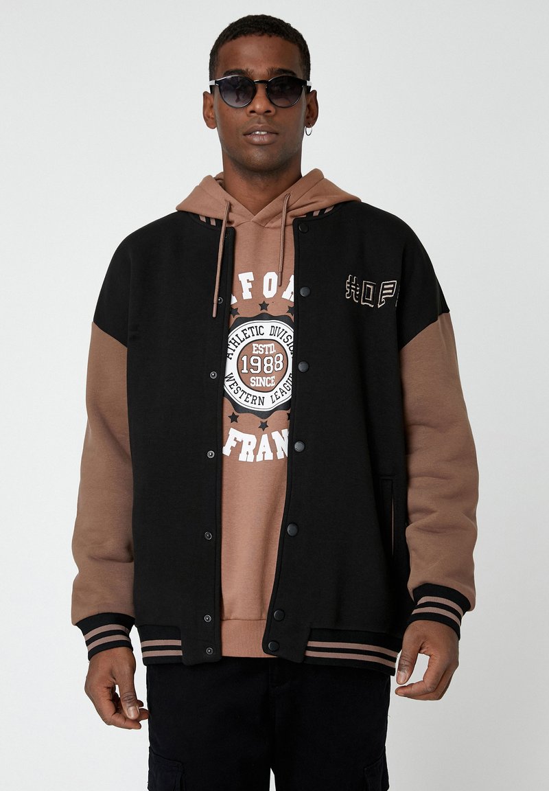 Chaqueta varsity negra y marrón con botones de presión, puños a rayas y bolsillos laterales sobre un sudadera beige con texto gráfico y estrellas.