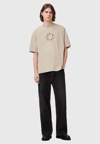 Beige, oversized póló, kerek nyakkivágással, elöl kör alakú fekete grafikával, laza fekete farmernadrággal és fekete cipővel párosítva.