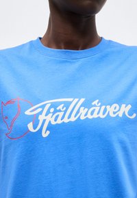 Άτομο που φοράει μπλε πουκάμισο με λευκή καλλιγραφική επιγραφή "Fjällräven" και κόκκινο λογότυπο κεφαλής αλεπούς στο στήθος.