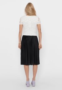 Maglietta bianca in pizzo a maniche corte abbinata a una gonna nera a pieghe, caratterizzata da una texture liscia. L'outfit è completato da scarpe con tacco a blocco color lilla.