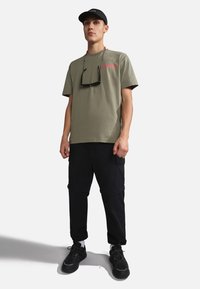 Camiseta de algodón verde oliva con texto rojo, combinada con pantalones cargo negros, zapatillas negras y una gorra negra. Gafas de sol colgando del cuello.