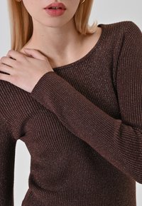 Femme portant un haut à manches longues côtelé marron avec des fils métalliques, la main reposant sur son épaule opposée, sur fond clair.
