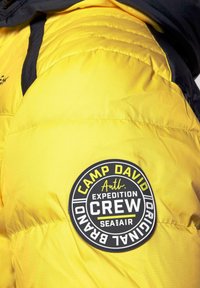 Żółta kurtka puchowa z czarnym kapturem, pikowaną teksturą oraz okrągłą czarno-białą łatą z napisem "CAMP DAVID EXPEDITION CREW."