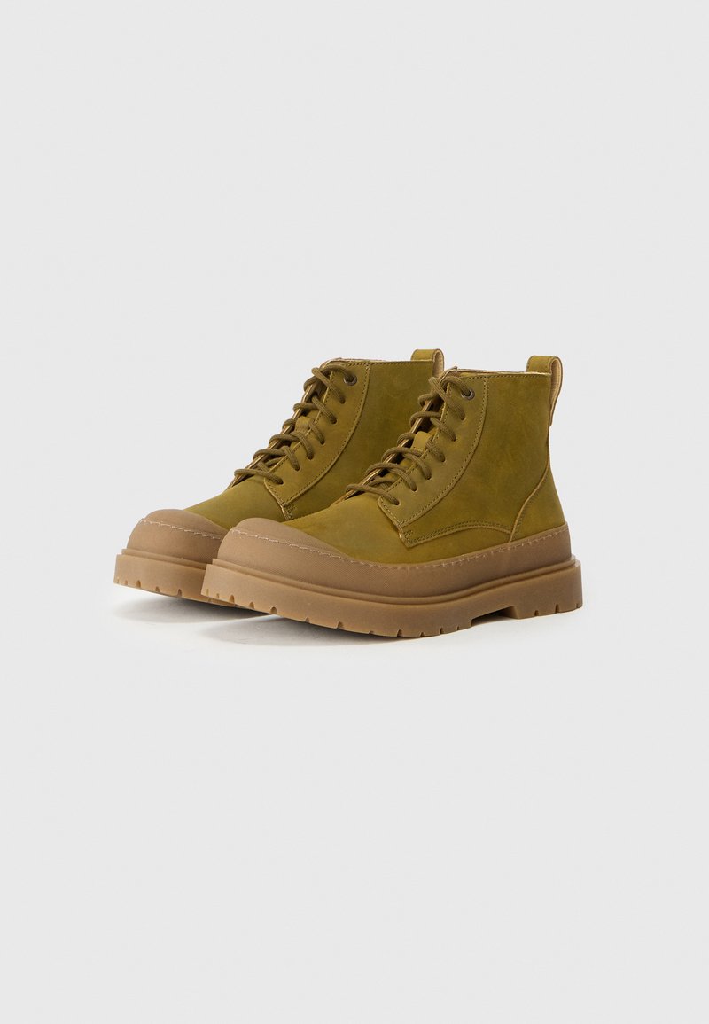 Bottes en suède vert olive à la cheville avec semelle en caoutchouc beige, bout rond et système de laçage par œillets. Design simple avec une languette à l'arrière.