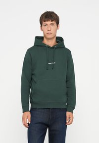 Haut à capuche vert foncé en tissu doux avec une poche avant, un cordon de serrage pour la capuche, et un logo brodé blanc sur la poitrine.