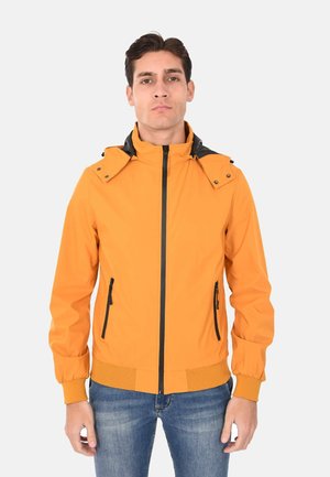 Uomo con giacca gialla senape con cerniera nera e dettagli, e jeans blu, in piedi contro uno sfondo chiaro e uniforme.