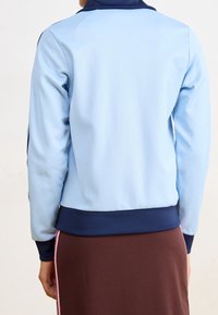 Osoba v světle modré bundě s tmavě modrými manžetami a límcem, v kombinaci s hnědými sukněmi s růžovým bočním pruhem, zobrazená zezadu.