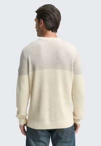 Crème ribgebreide sweater met een grijze bovenkant, voorzien van een ronde hals, lange mouwen en een gestructureerde brei. Gedragen met een blauwe spijkerbroek.