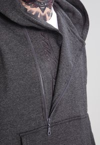 Szary rozpinany kaptur wykonany z miękkiego materiału, z podszewką w kapturze, przednim zamkiem błyskawicznym z uchwytem oraz wytłoczonym wzorem na okolicy klatki piersiowej.