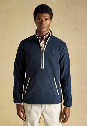 Mann trägt einen marineblauen Fleecesweater mit Viertelreißverschluss und cremefarbener Einfassung über einem rot karierten Hemd und hellbeigen Hosen und steht vor einem neutralen Hintergrund.