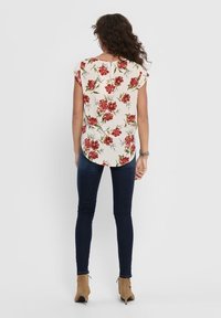 Blusa floreale con fiori rossi su uno sfondo crema, maniche corte, orlo arrotondato e dettaglio a occhiello sul retro. Abbinata a jeans scuri e stivali alla caviglia.