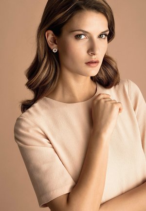 Frau mit schulterlangem braunem Haar trägt beige kurzärmliges Oberteil und runde silberne Ohrringe, hält ihre Hand nahe an der Brust vor braunem Hintergrund.