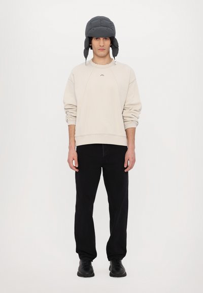 Pull beige avec des manches à revers, pantalon noir et un chapeau de trappeur gris matelassé avec des rabats d'oreilles. Le modèle se tient contre un fond blanc.