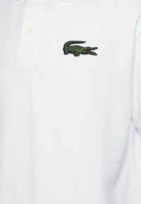 Valkoinen puuvillapaita, jossa on vasemmassa rinnassa vihreä brodeerattu krokotiililogo, klassinen kaulus ja kaksi nappia.