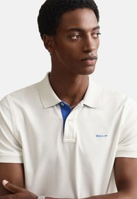 Polo majica v svetli krem barvi z modrim ovratnikom. Mehka tekstura, trije gumbi na sprednji strani in majhen modri logotip na prsih.