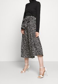 Kvinna som bär en svart långärmad polotröja, svart midikjol med vitt prickmönster och knytning i midjan, som kombineras med vita högklackade sandaler med ankelsnöre.