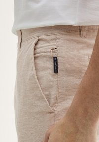 Pantalones de lino beige con un bolsillo lateral y detalles de cremallera. La tela tiene un aspecto texturizado, con una pequeña etiqueta de marca.
