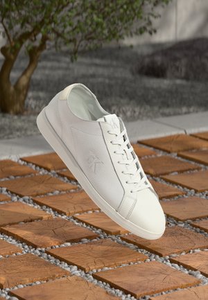 Biały skórzany sneaker z sznurowadłami unoszący się nad drewnianą płytką i żwirową powierzchnią, w tle rozmyte małe zielone drzewo.