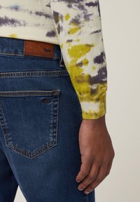 Krema, rumena in vijolična majica s dolgimi rokavi v tie-dye stilu, kombinirana s temno modrimi denim hlačami, ki imajo rjavo usnjeno oznako in šivano podrobnost na žepu.