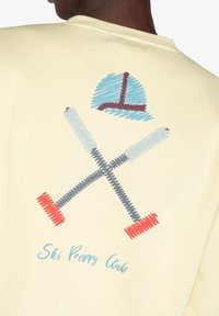Sudadera de color amarillo claro con un diseño gráfico de esquís cruzados y un gorro en azul, rojo y gris, con el texto "Ski Preppy Club."