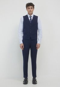 Un giovane uomo sta in piedi di fronte, indossando un gilet a righe navy, pantaloni abbinati, una camicia a strisce blu chiaro, una cravatta a righe e scarpe nere eleganti.